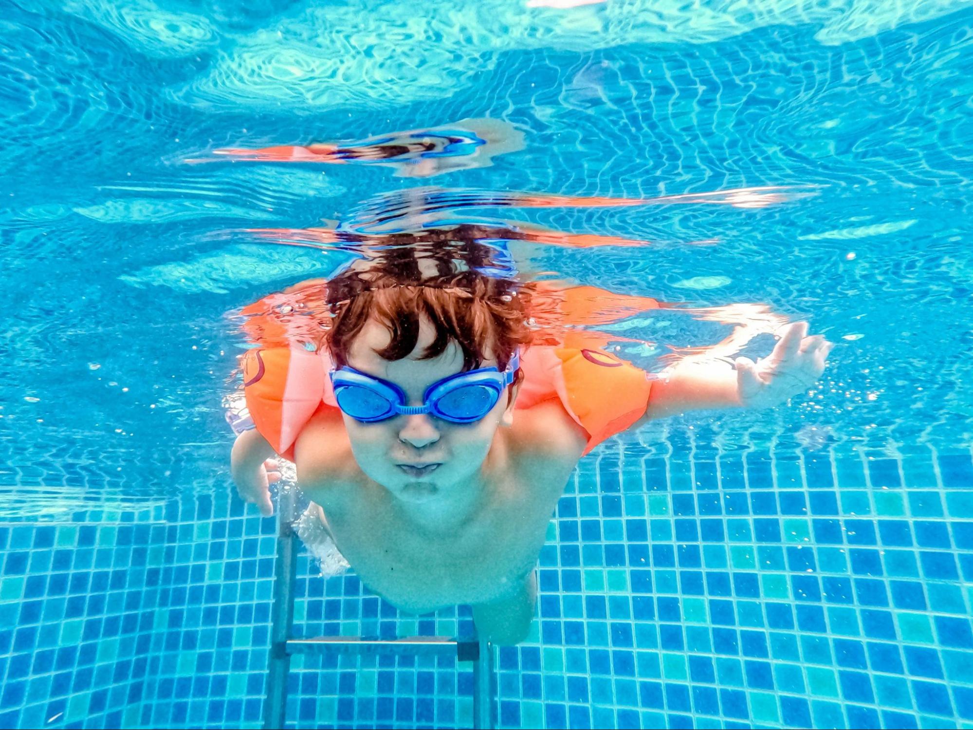 A young boy in goggles and arm floaties is actively swimming underwater, an action that demonstrates the water confidence built by the Best Kids Swimming Lessons In Singapore.